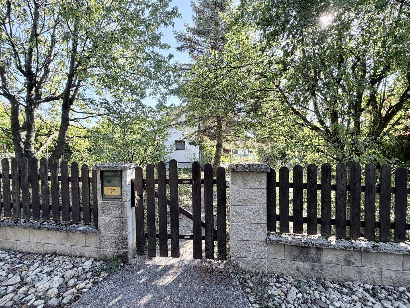 Rustikaler Holzzaun mit Steinsäulen und Eingangstor zum Grundstück, umgeben von Bäumen.