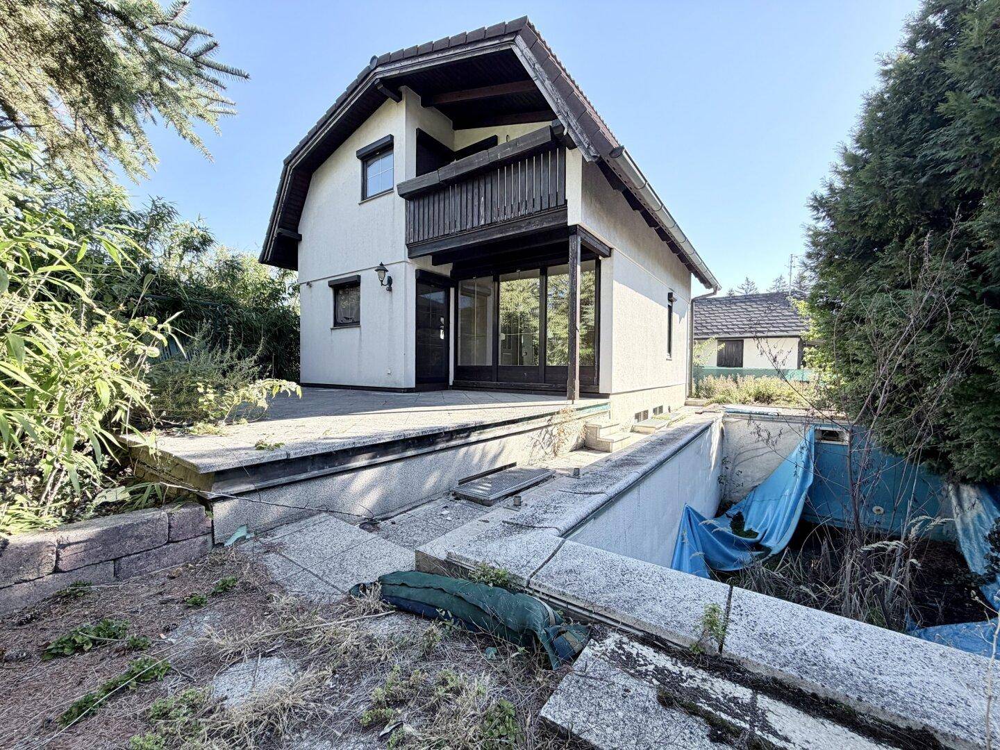 Hausansicht mit Terrasse und einem alten, renovierungsbedürftigen Poolbereich im Garten.