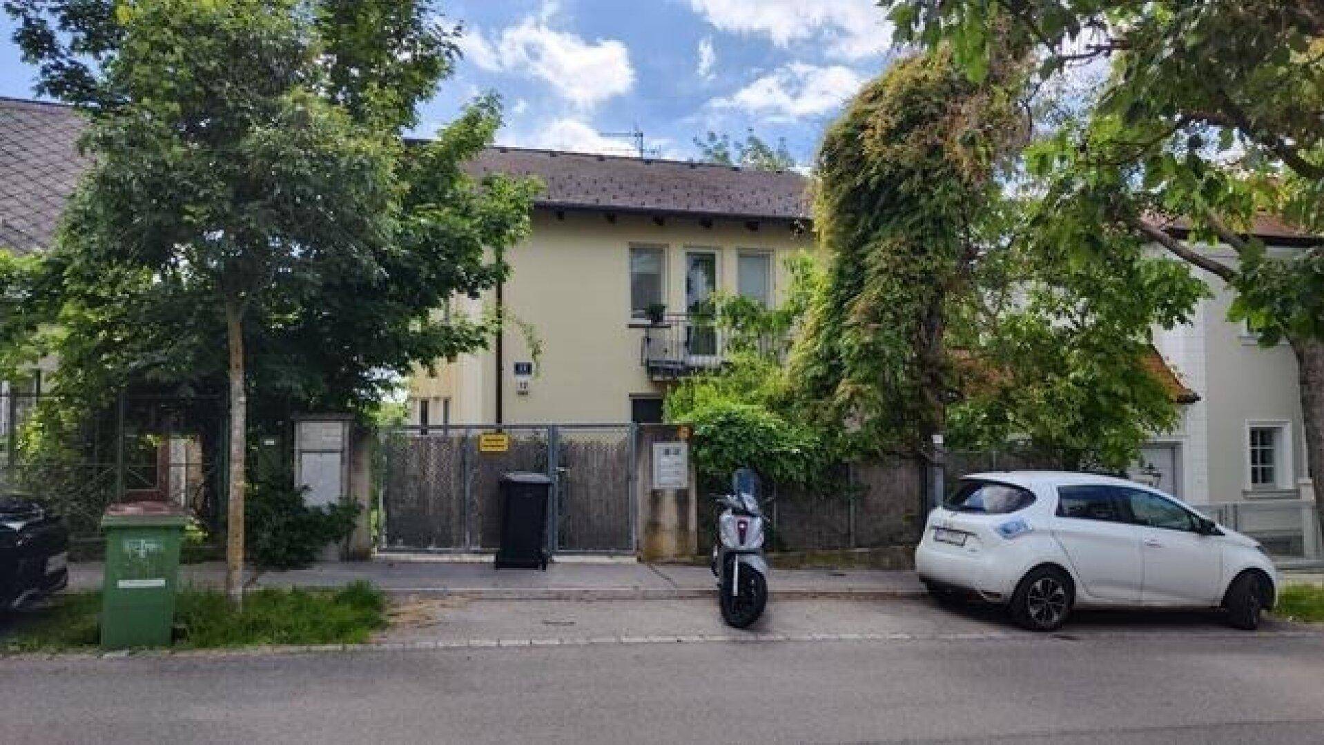 Ansicht des Hauses von der Straße mit Zaun, Bäumen und geparkten Fahrzeugen davor.