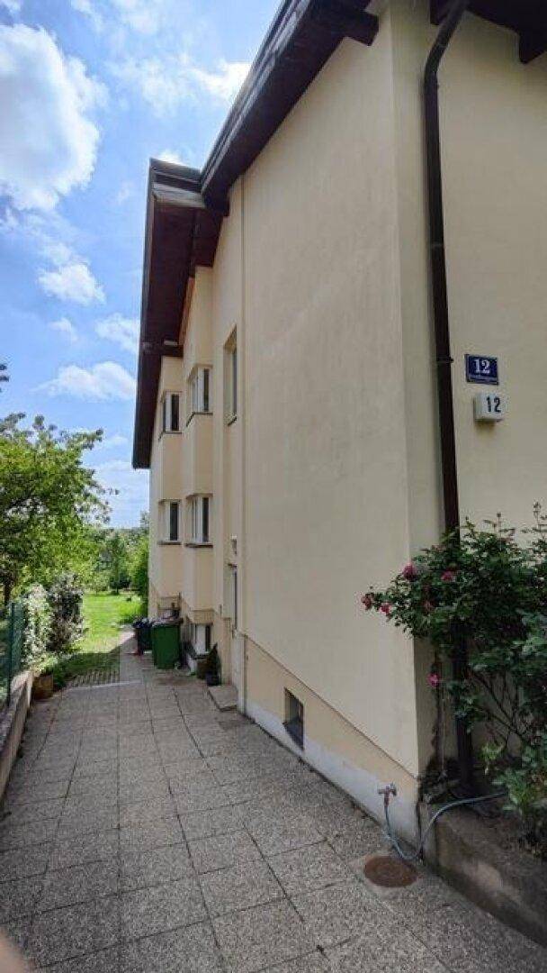 Seitenansicht des Hauses mit gepflastertem Weg, Fenstern und der Hausnummer 12.