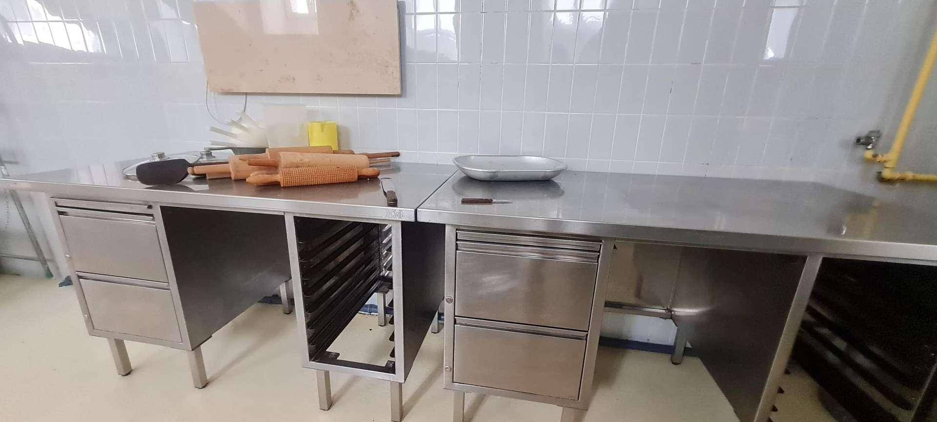 Detailansicht von zwei Edelstahl-Arbeitstischen mit Utensilien in einer professionellen Küche mit weißen Fliesen.