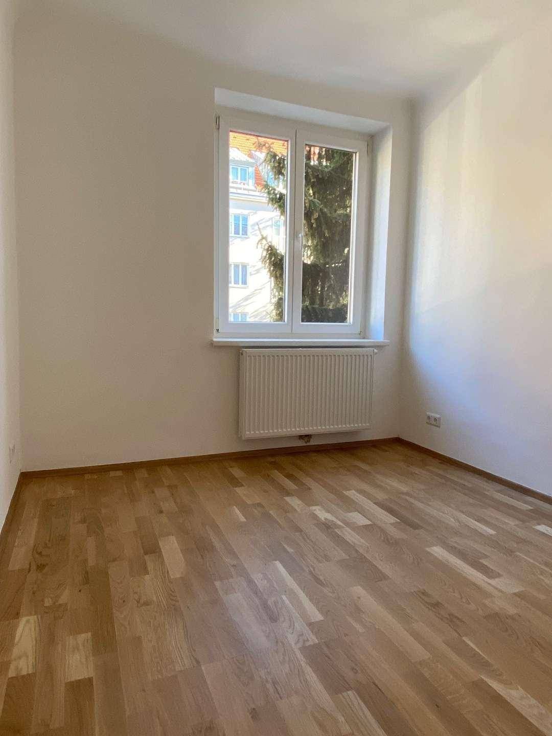 Helles, leeres Zimmer mit Parkettboden, einem großen Fenster und Heizkörper, bereit zur Gestaltung.