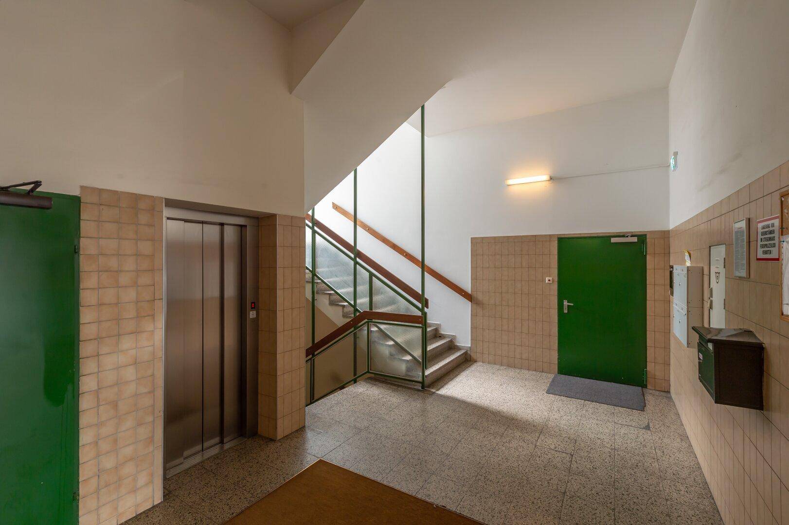 Eingangsbereich mit Aufzug, Treppenhaus und gefliesten Wänden in einem funktionalen Stil.