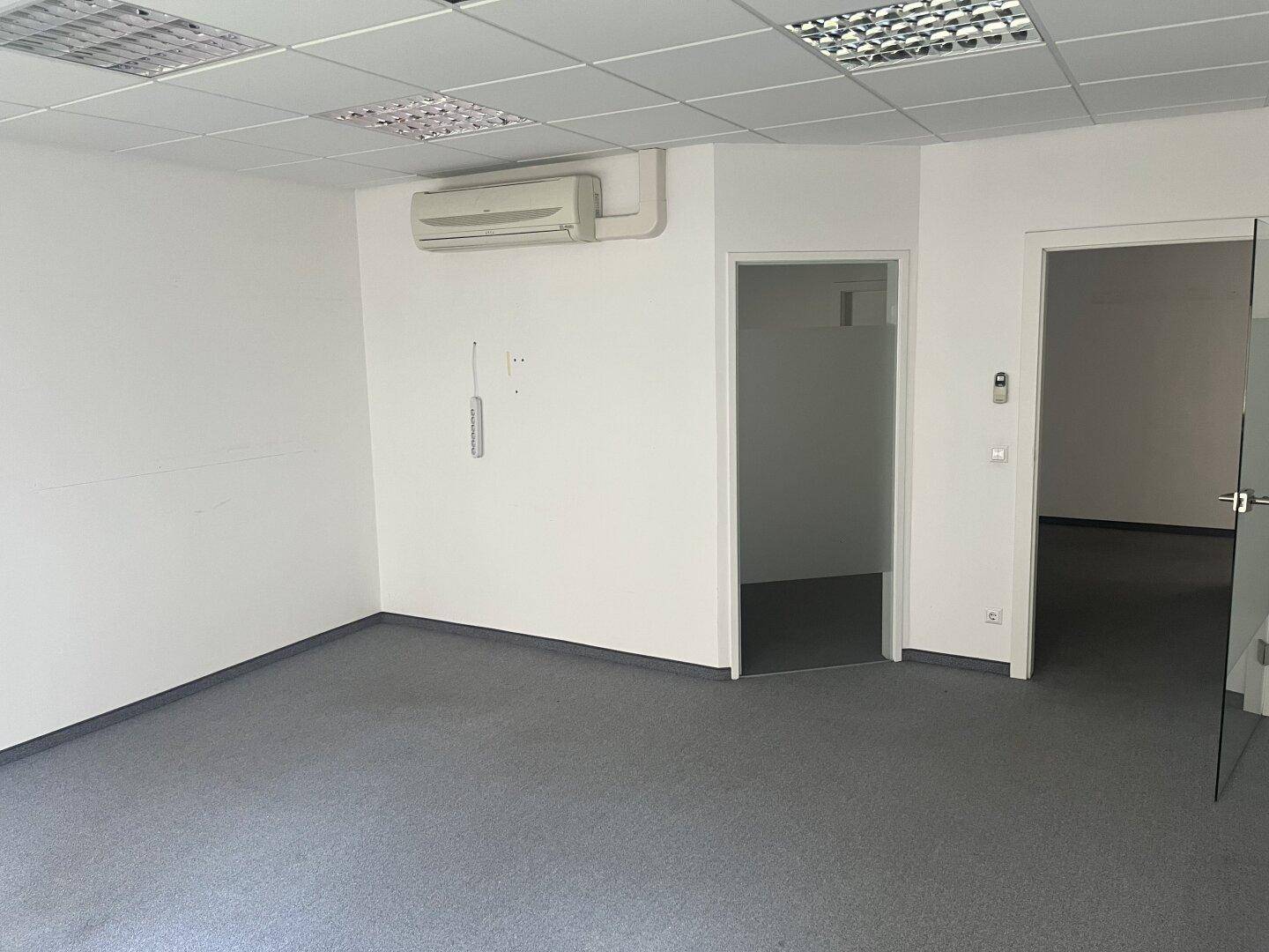 Helles Büro mit Teppichboden, Klimaanlage und einer Tür, die zu einem weiteren Raum führt.