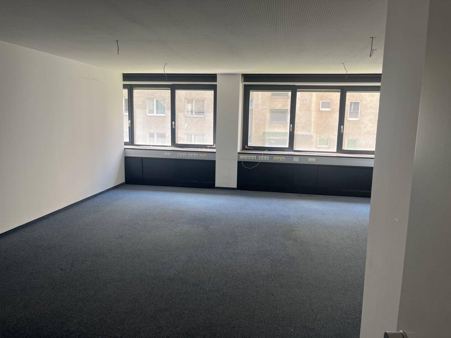 Helles Büro mit blauem Teppichboden, großen Fenstern und integrierten Wandablagen.