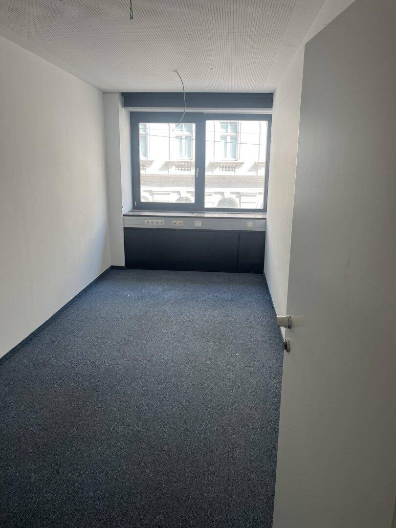 Kleines Büro mit blauem Teppichboden, einem großen Fenster und integrierten Wandablagen.