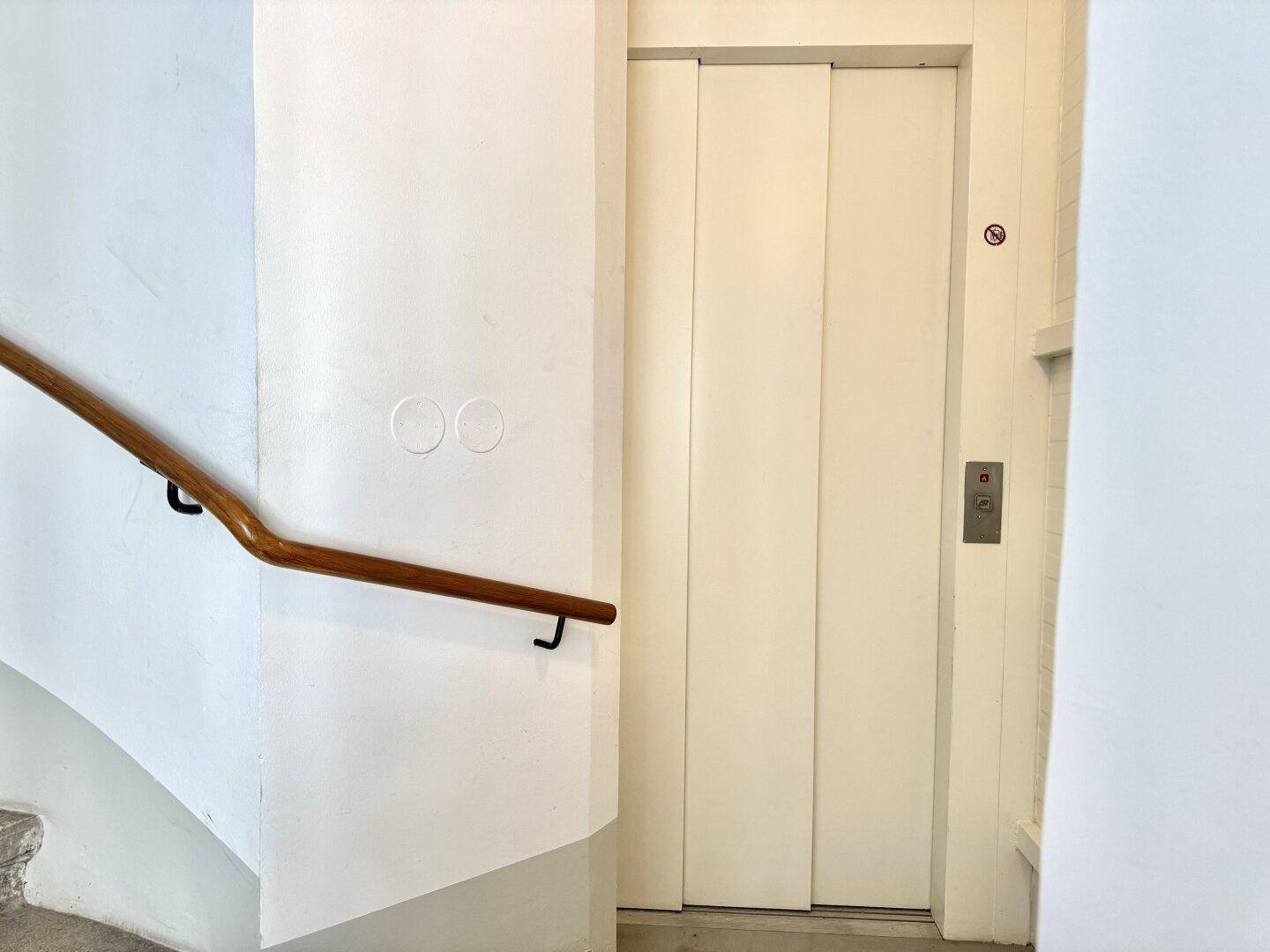 Aufzugstür in einem hellen Treppenhaus mit Holzhandlauf, der zu den oberen Etagen führt.