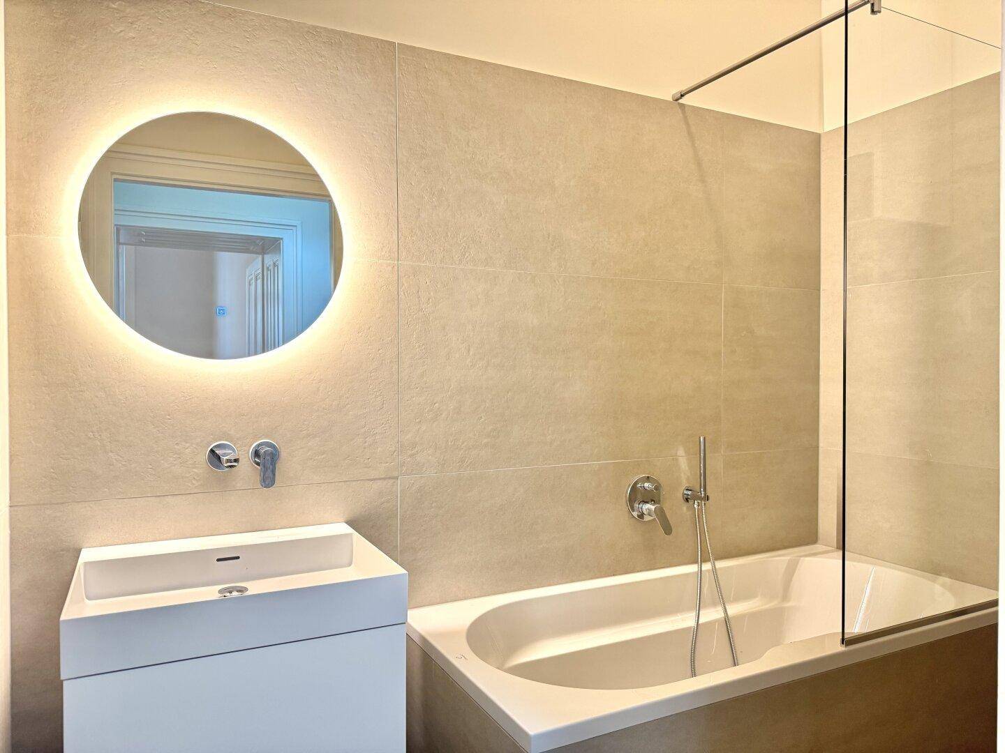 Modernes Badezimmer mit Badewanne, elegantem Waschtisch und beleuchtetem Rundspiegel.