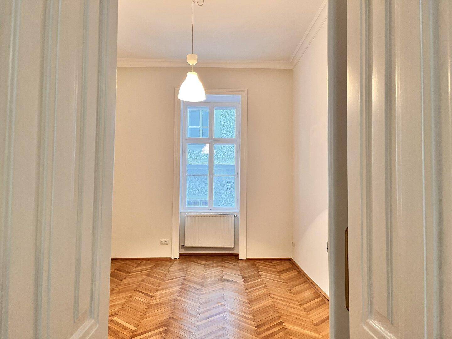 Freundliches Zimmer mit klassischem Fischgrätparkett und Fenster, zugänglich durch eine Flügeltür.