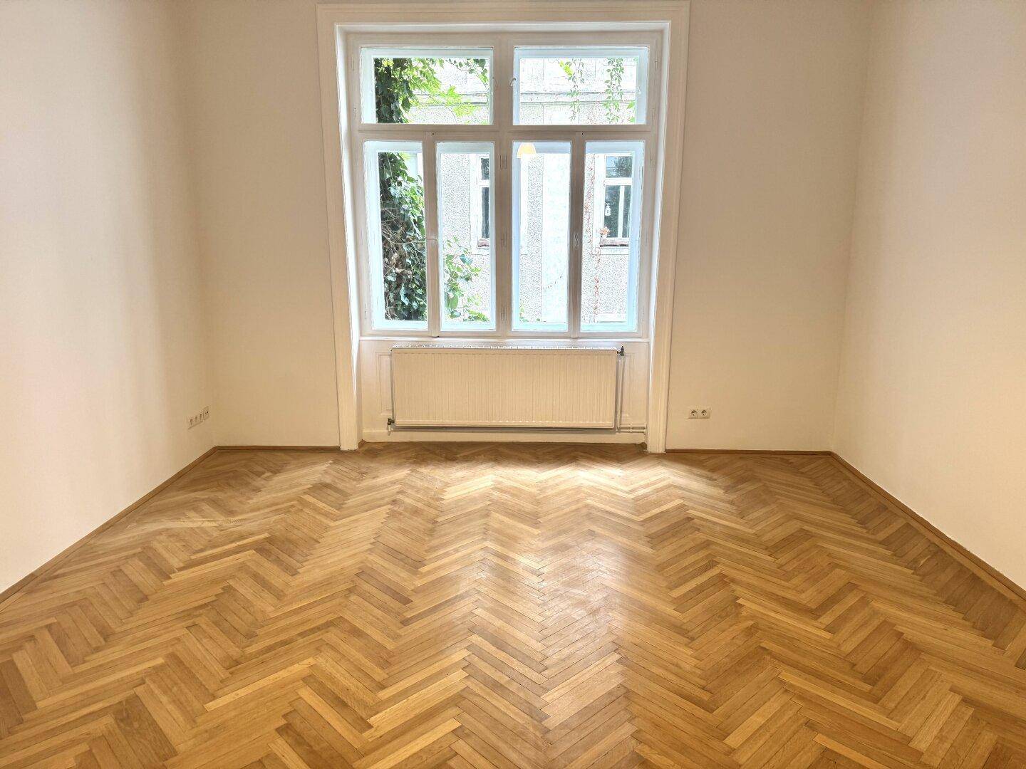 Geräumiges Zimmer mit Fischgrätparkett und großem Fenster, ideal für Wohn- oder Schlafbereich.