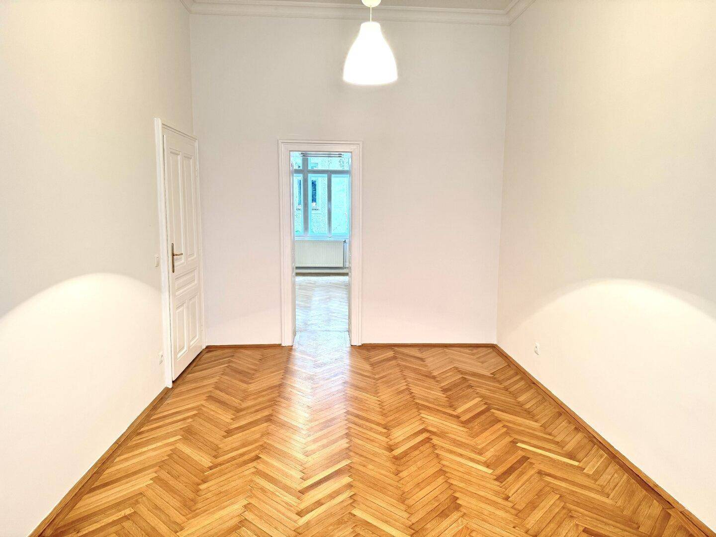 Geräumiges Zimmer mit Fischgrätparkett und Blick in einen weiteren Raum, ideal für offenes Wohnen.