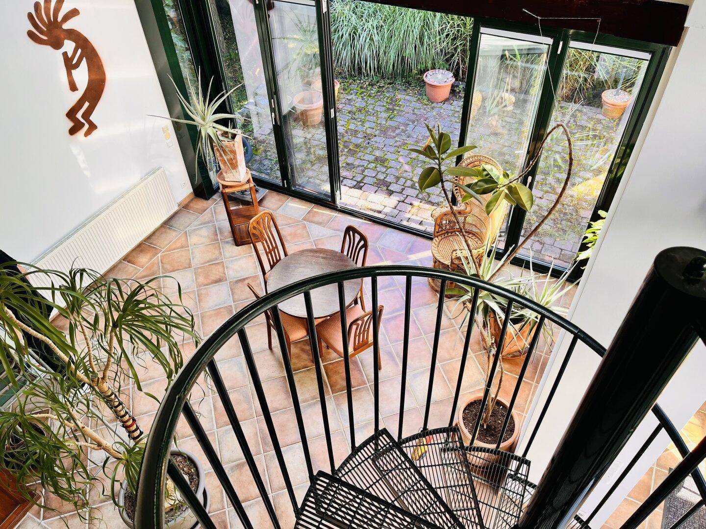 Wendeltreppe führt hinunter in den hellen Wintergarten mit Fliesenboden und Pflanzen.