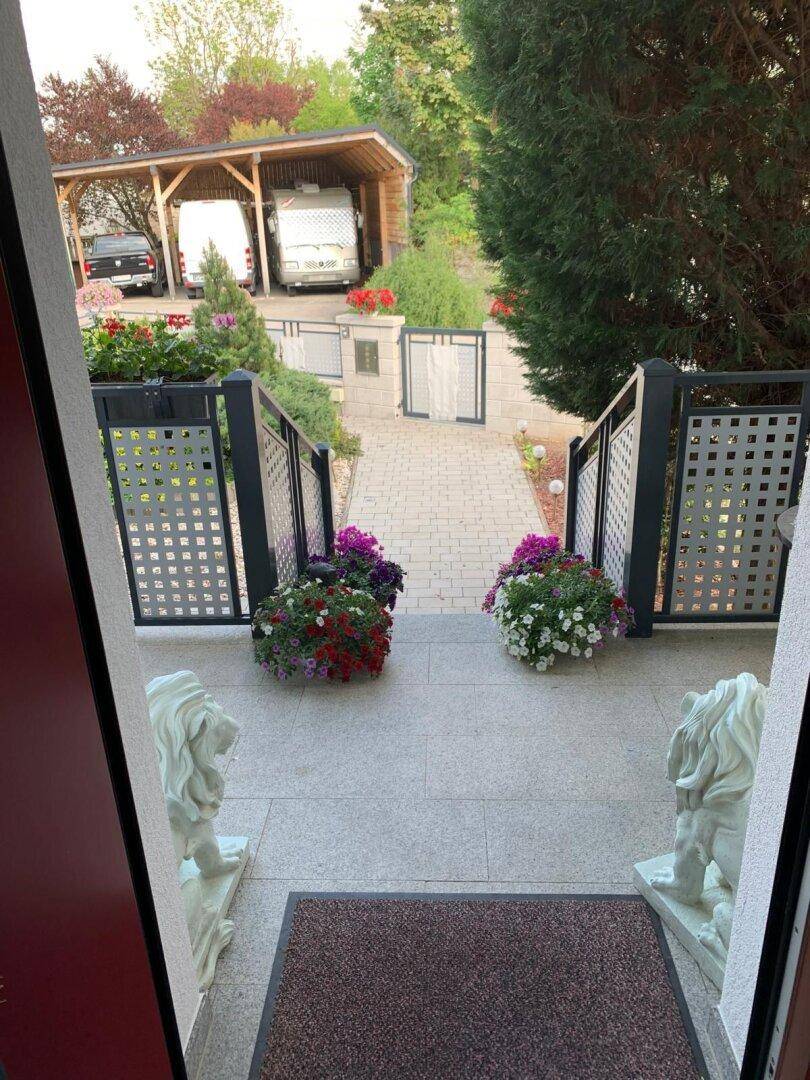 Blick vom Hauseingang über einen gepflasterten Weg zu einem Carport im Hintergrund.
