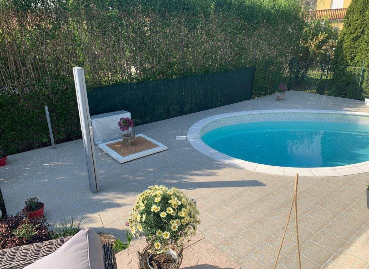 Ovaler Swimmingpool umgeben von einer hell gefliesten Terrasse und einer grünen Hecke.