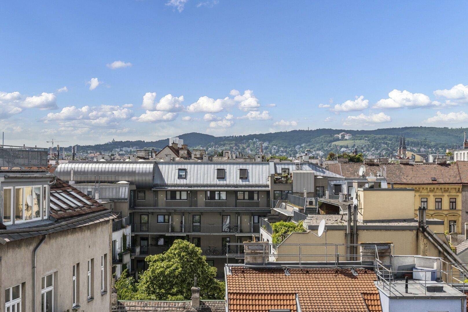Weitläufiger Blick über die städtische Dachlandschaft mit verschiedenen Gebäudetypen und grünen Hügeln im Hintergrund.
