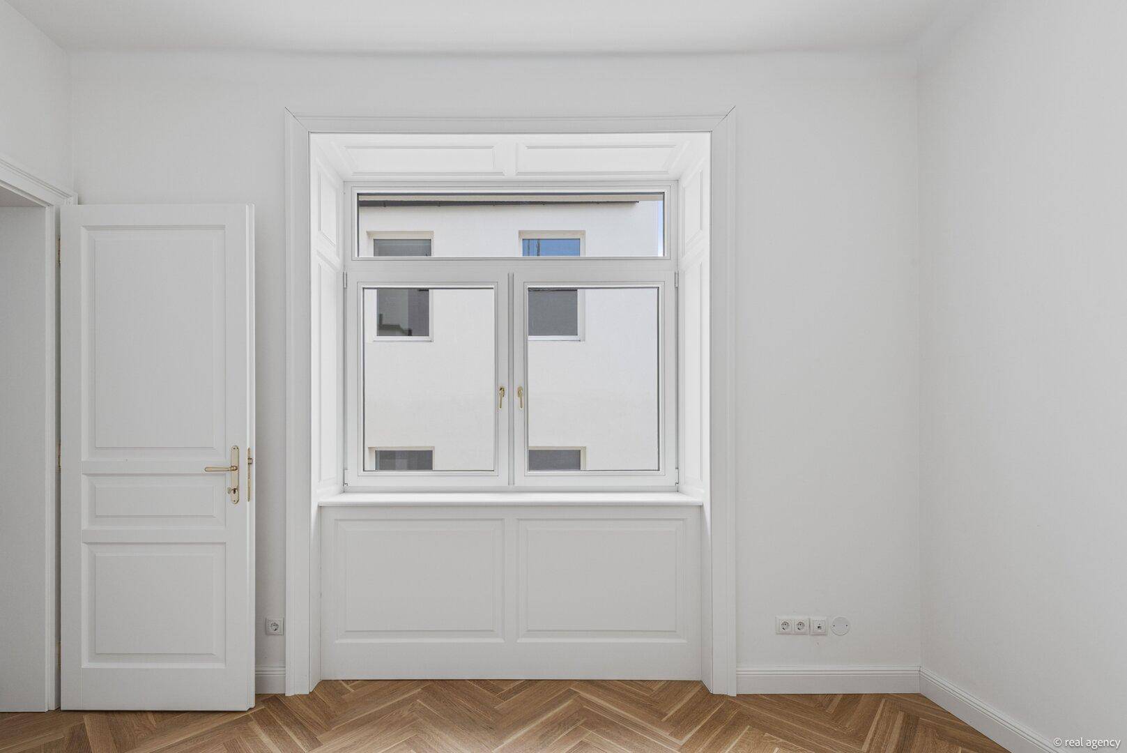Helles Zimmer mit Fischgrätparkett und Fenster mit Blick auf die gegenüberliegende Fassade.