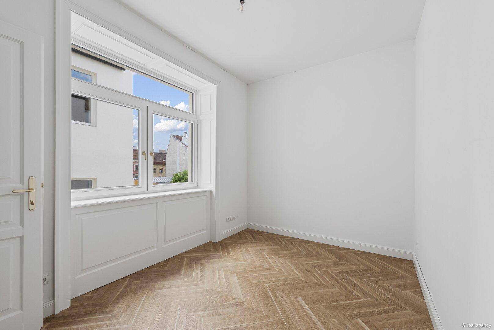 Helles Schlafzimmer mit Fischgrätparkett und Fenster mit Blick auf die Nachbargebäude.