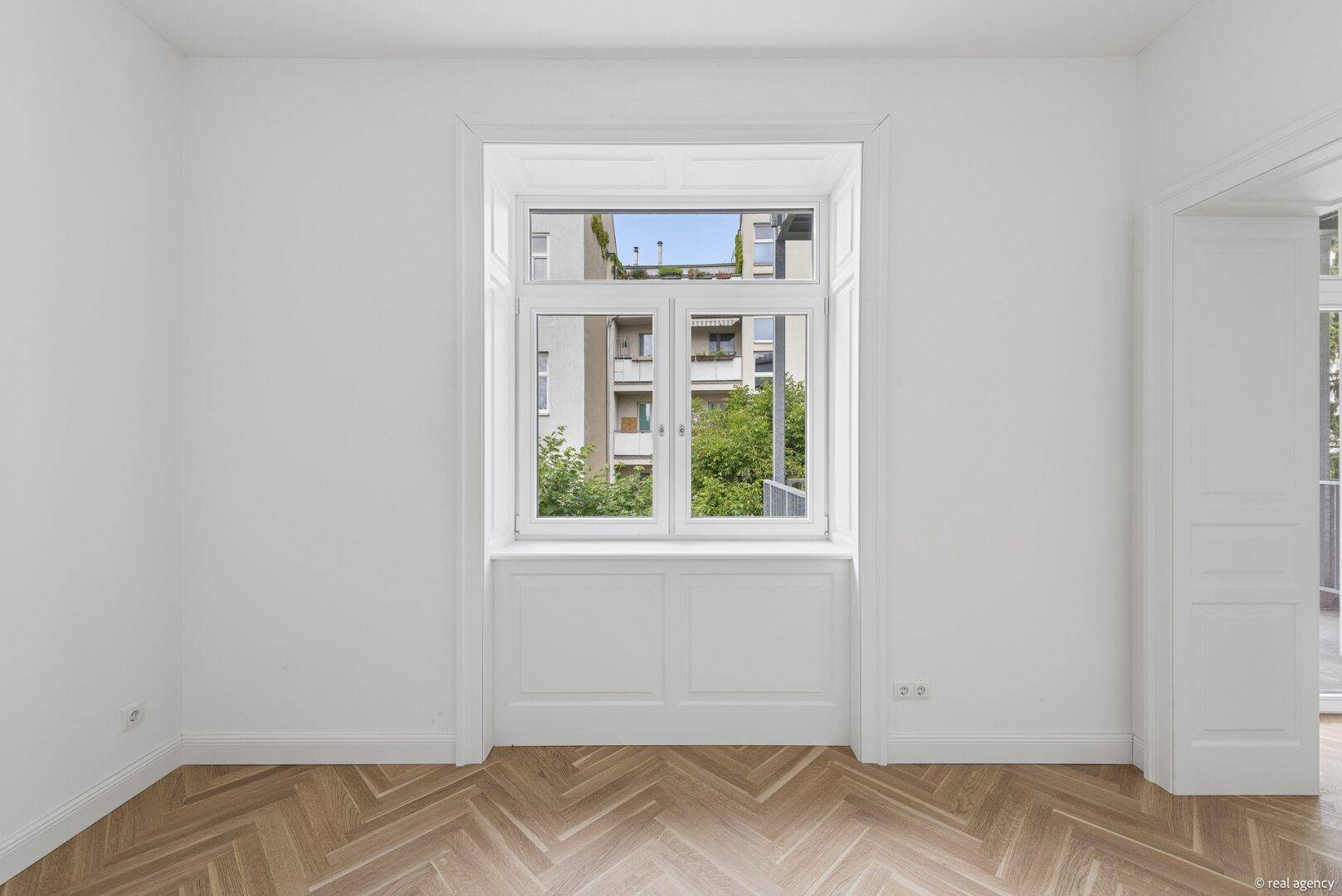 Helles, leeres Zimmer mit Fischgrätparkett und großem Fenster mit Blick ins Grüne.