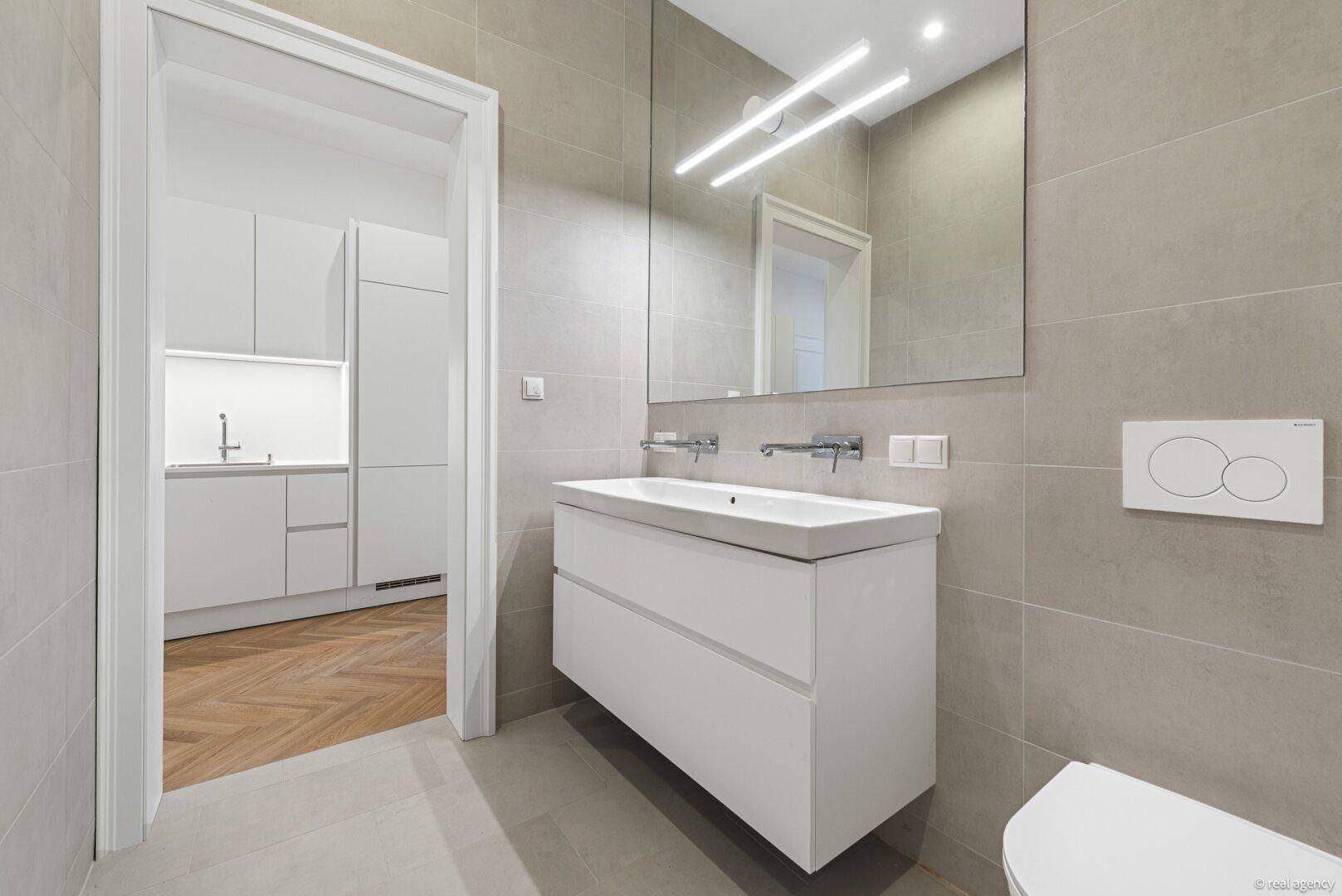 Elegantes Badezimmer mit großem Spiegel, modernem Waschtisch und Blick in die angrenzende Küche.