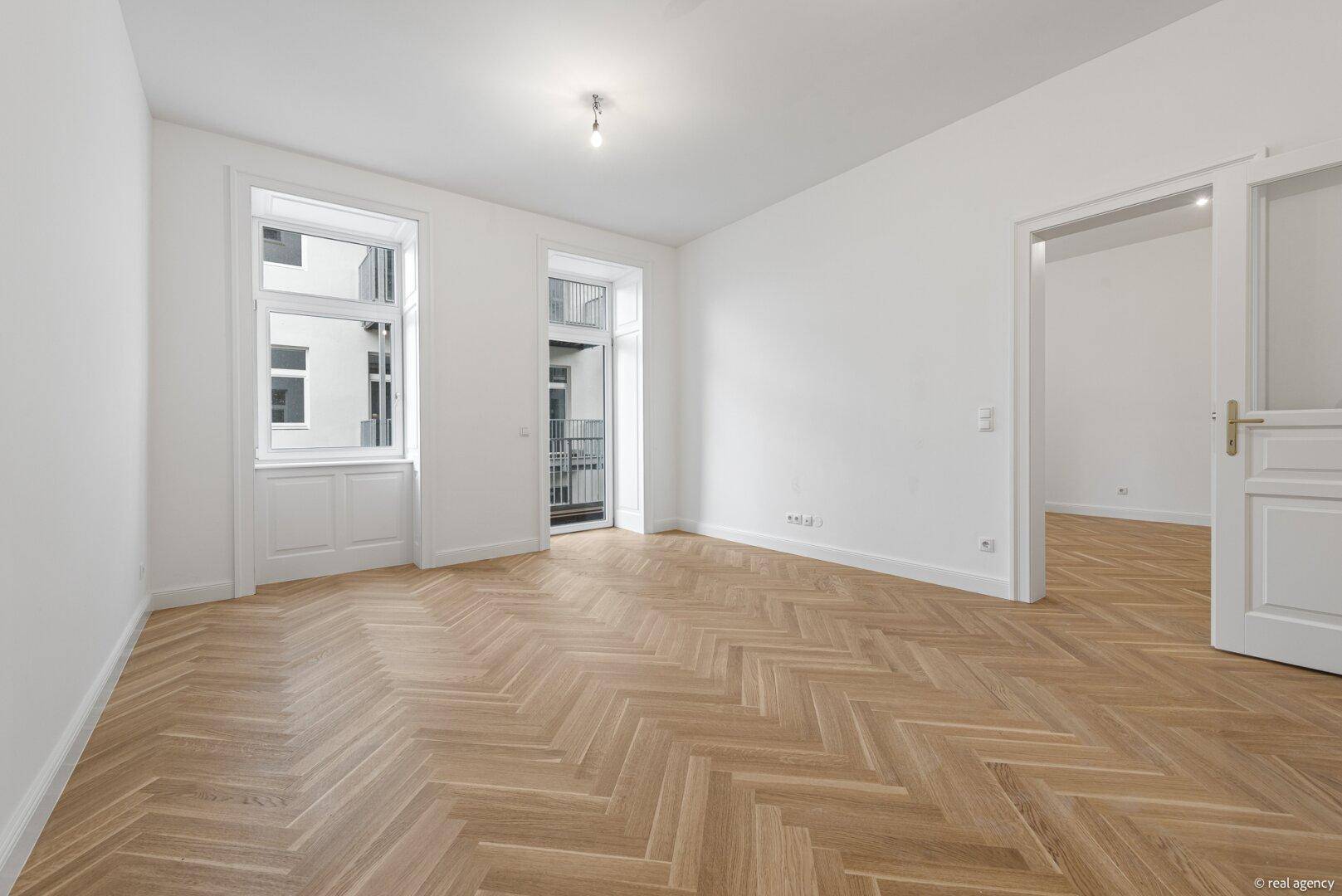 Geräumiges Wohnzimmer mit Fischgrätparkett, hohen Decken und Zugang zu einem französischen Balkon.