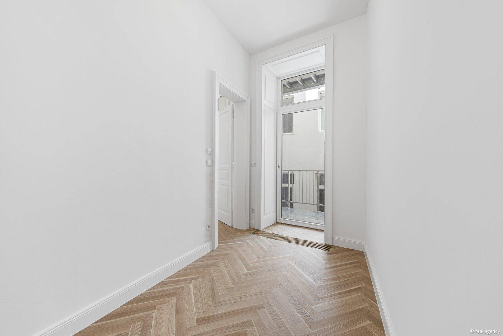 Lichtdurchfluteter Flur mit Fischgrätparkett und einem französischen Balkon am Ende.