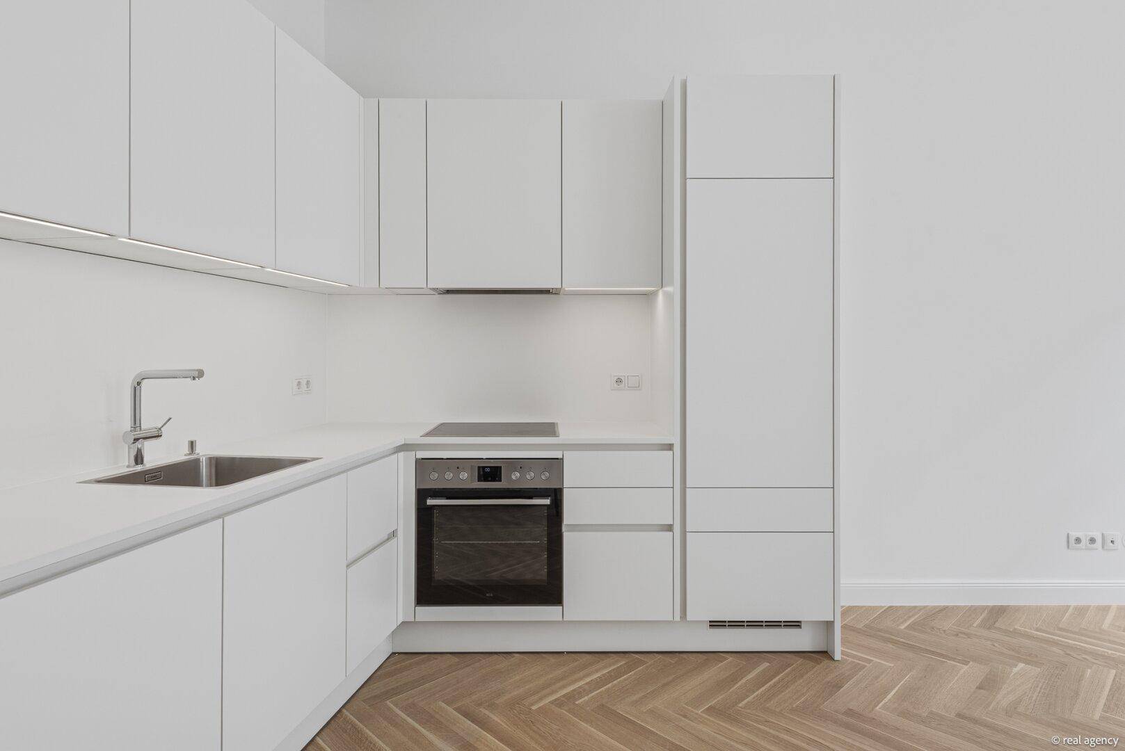 Moderne weiße Einbauküche mit Spüle, Backofen und Induktionskochfeld in einem hellen Raum.