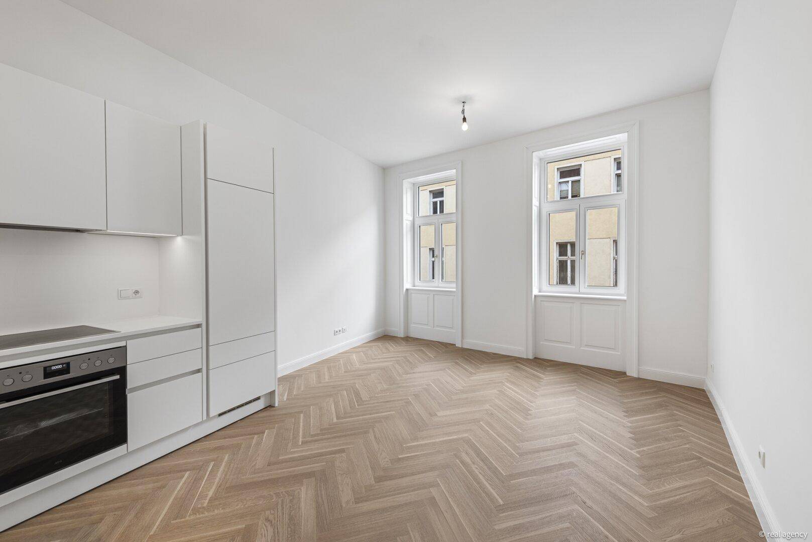Heller Wohnraum mit Fischgrätparkett und integrierter weißer Küchenzeile, zwei großen Fenstern und Blick auf die Stadt.
