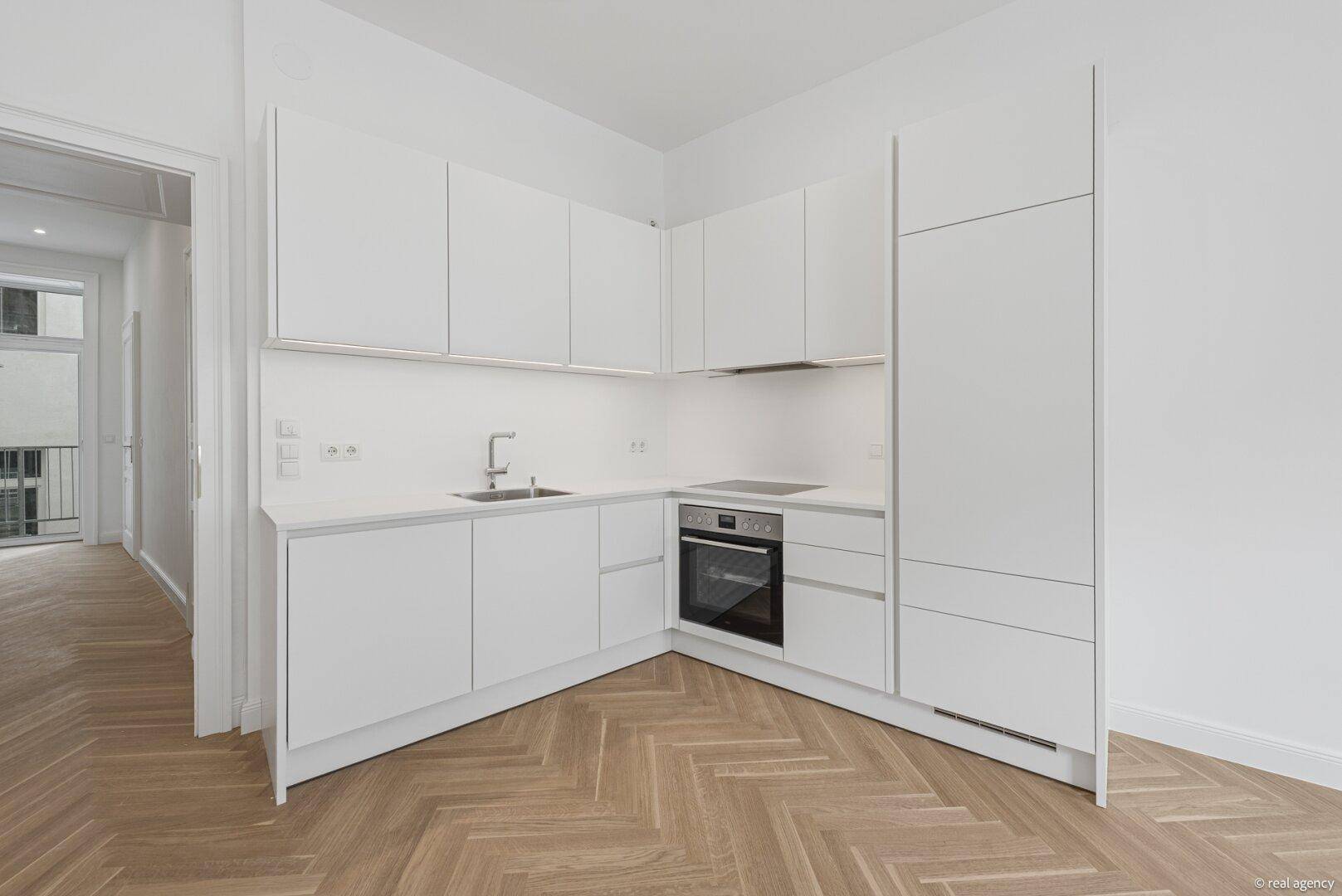 Moderne weiße Küchenzeile mit integrierten Geräten und Fischgrätparkett in einem hellen Raum.