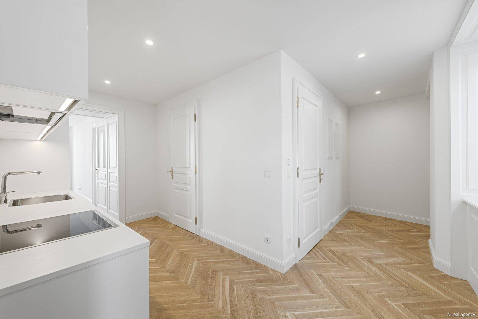 Helle Küche mit weißen Schränken und Parkettboden, angrenzend an weitere Zimmer.