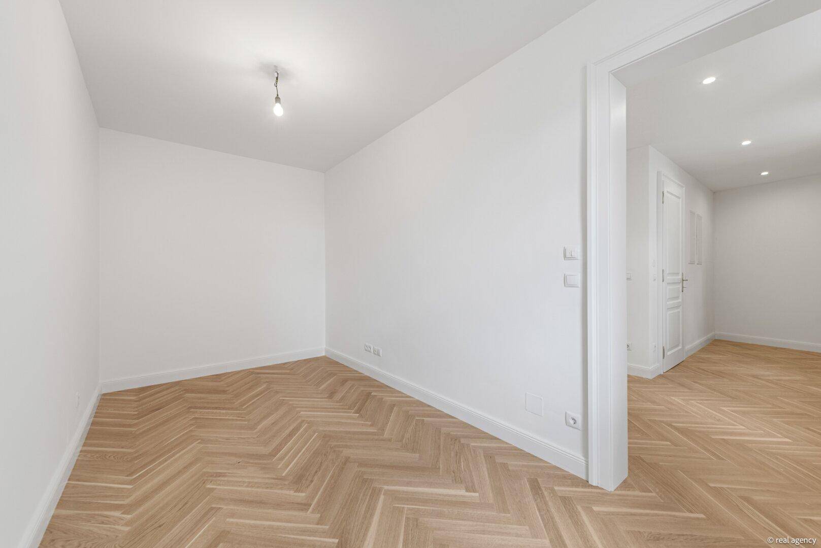 Helles Zimmer mit Fischgrätparkett und Zugang zu einem weiteren Raum.