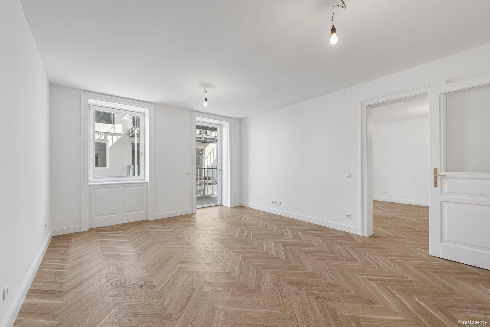 Helles Wohnzimmer mit Fischgrätparkett, großen Fenstern und Zugang zu einem Balkon.