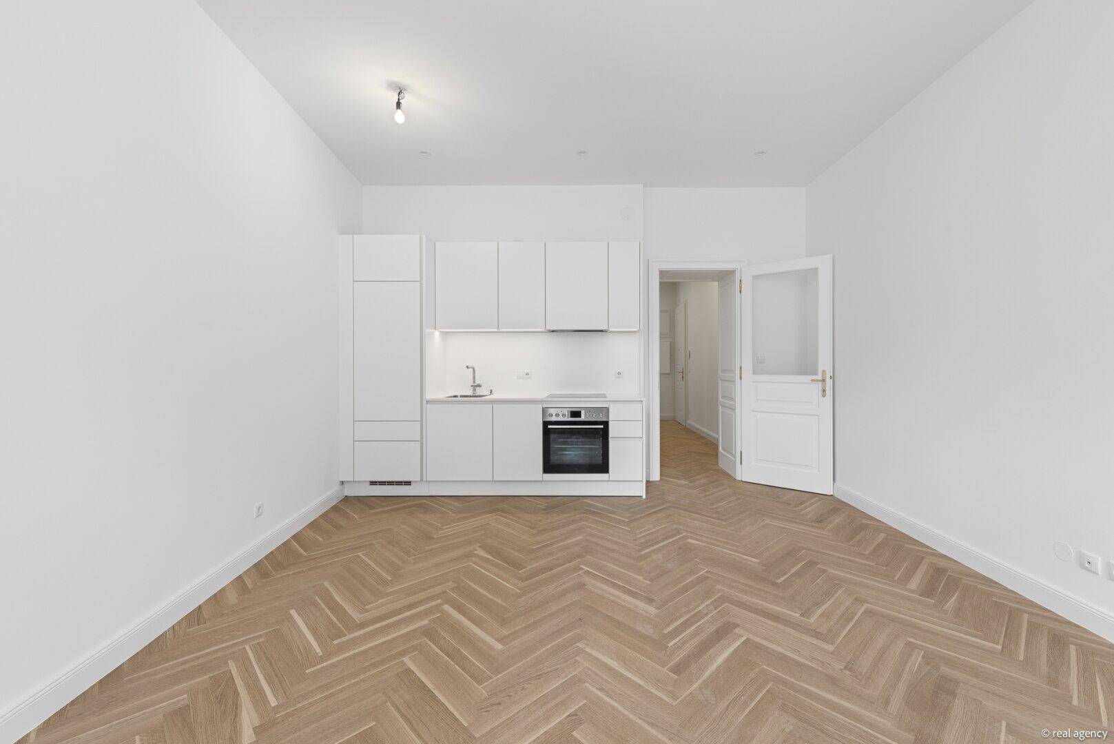 Offener Wohnbereich mit integrierter, moderner weißer Küche und elegantem Fischgrätparkett.