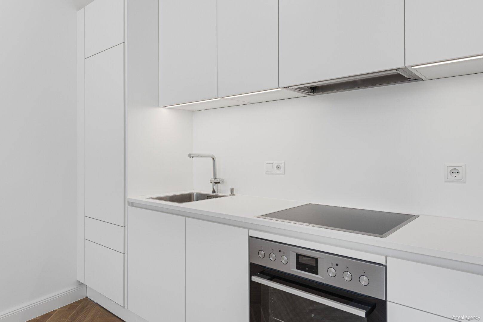 Moderne, weiße Einbauküche mit Induktionskochfeld, Backofen und integrierter Beleuchtung unter den Oberschränken.