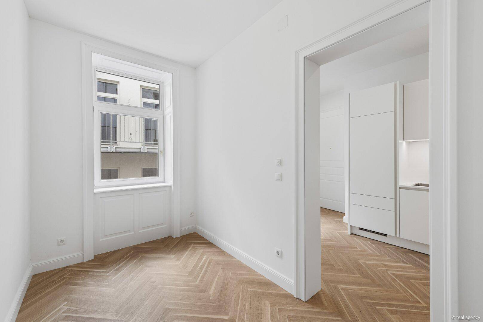 Heller Raum mit Fischgrätparkett und großem Fenster, offen zur angrenzenden Küche.
