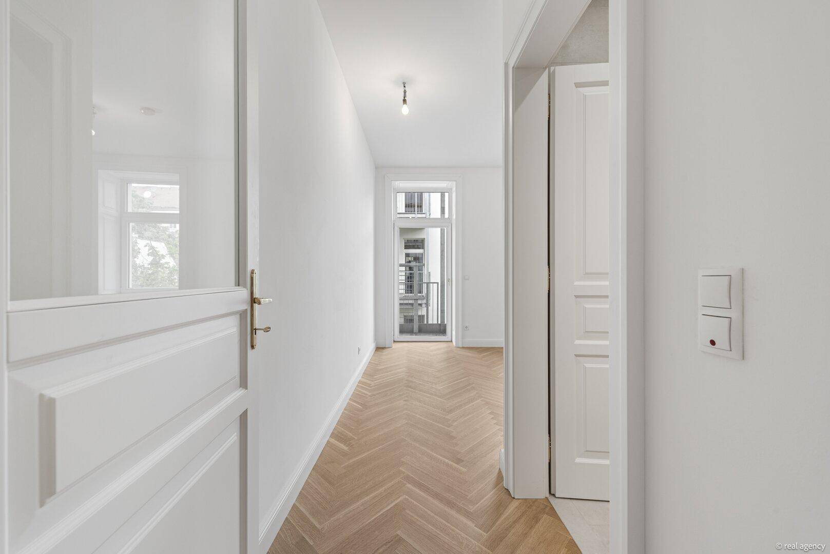 Langer, heller Flur mit Fischgrätparkett und weißen Türen, der zu einem Balkon führt.