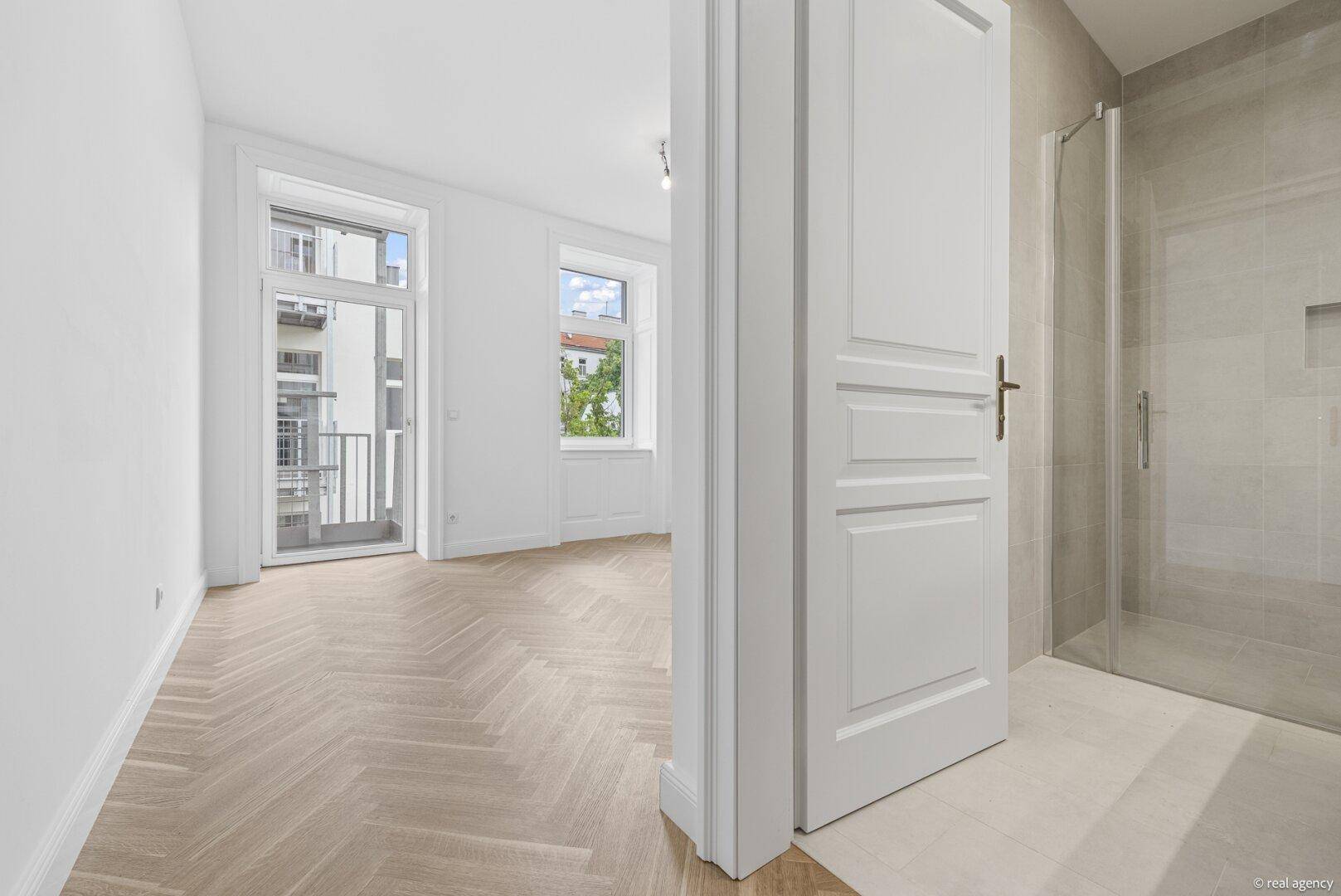 Heller Raum mit Fischgrätparkett und Balkonzugang, angrenzend an ein modernes Badezimmer.