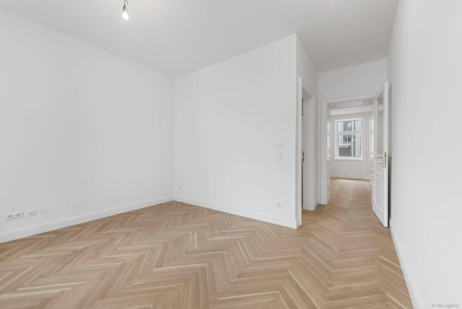 Geräumiger, heller Raum mit Fischgrätparkett und Zugang zu einem weiteren Bereich.