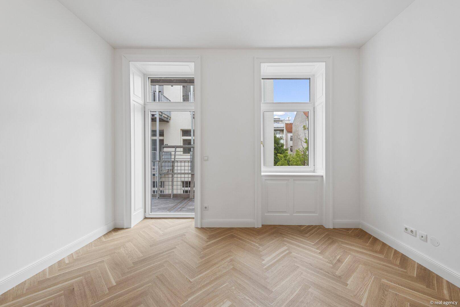 Lichtdurchfluteter Raum mit Fischgrätparkett, Balkontür und großem Fenster.