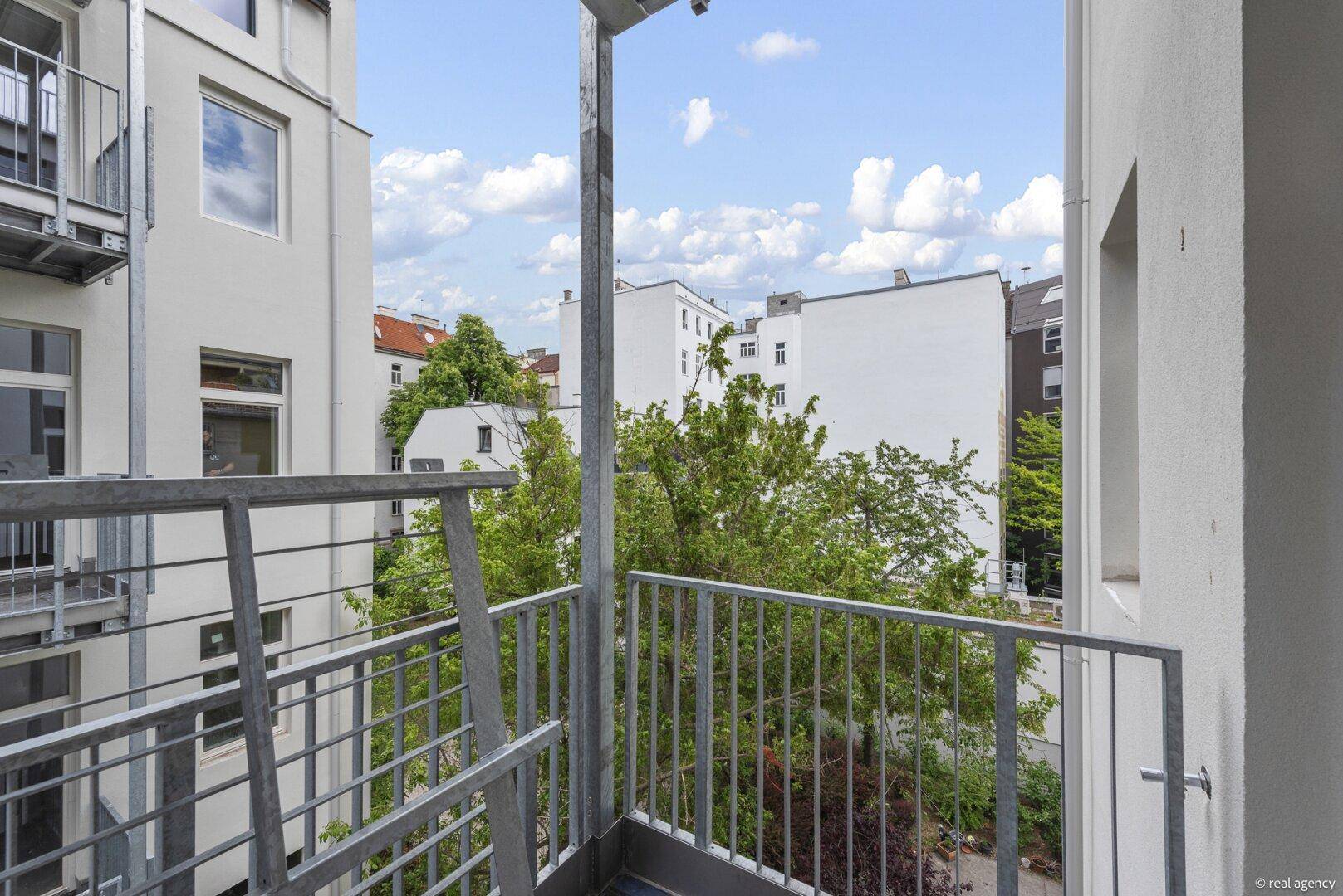 Kleiner Balkon mit Metallgeländer und Blick auf die umliegenden Gebäude und Bäume.