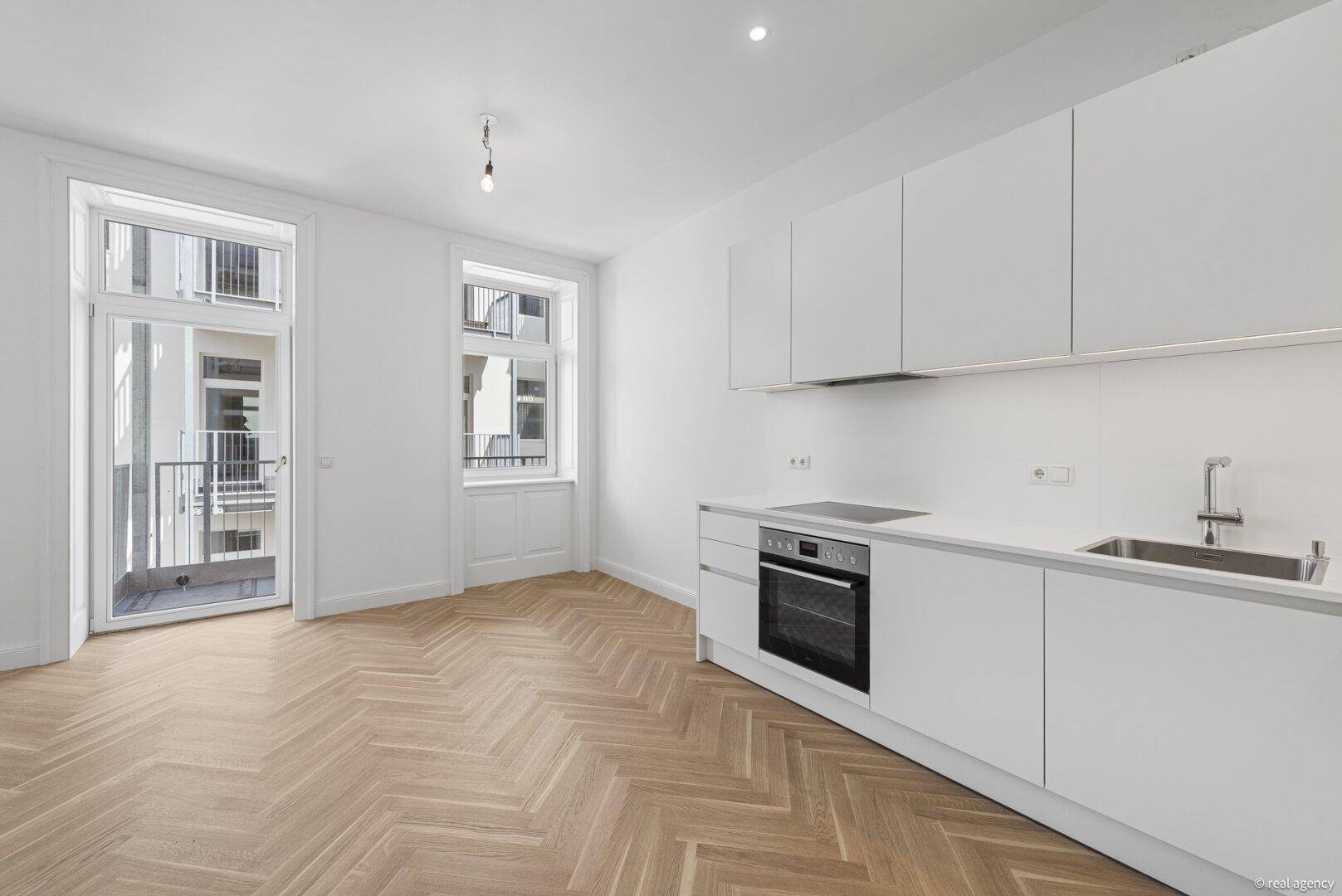 Helle offene Küche mit moderner Einbauküche, Fischgrätparkett und Zugang zu einem Balkon.