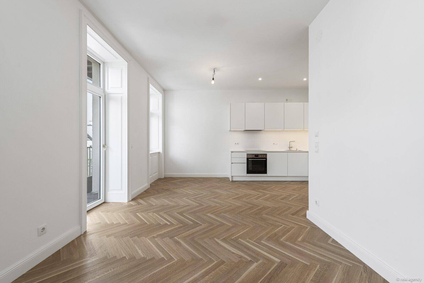 Geräumiger Wohnbereich mit Fischgrätparkett, offener Küche und Zugang zu einem Balkon.