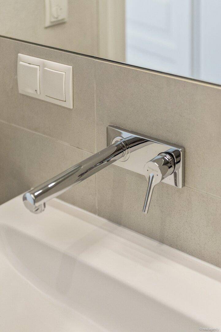 Wandmontierter Wasserhahn über einem Waschbecken mit grauen Fliesen im Badezimmer.