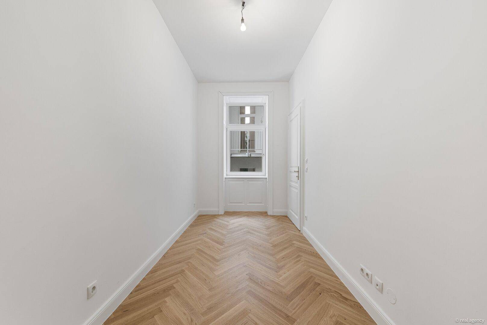 Langer, heller Raum mit Fischgrätparkett und Fenster, ideal als Schlaf- oder Arbeitszimmer.