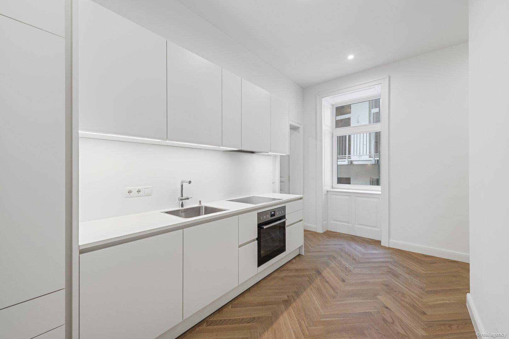 Moderne Einbauküche mit weißen Fronten, integriertem Kochfeld und Ofen, sowie Fenster.