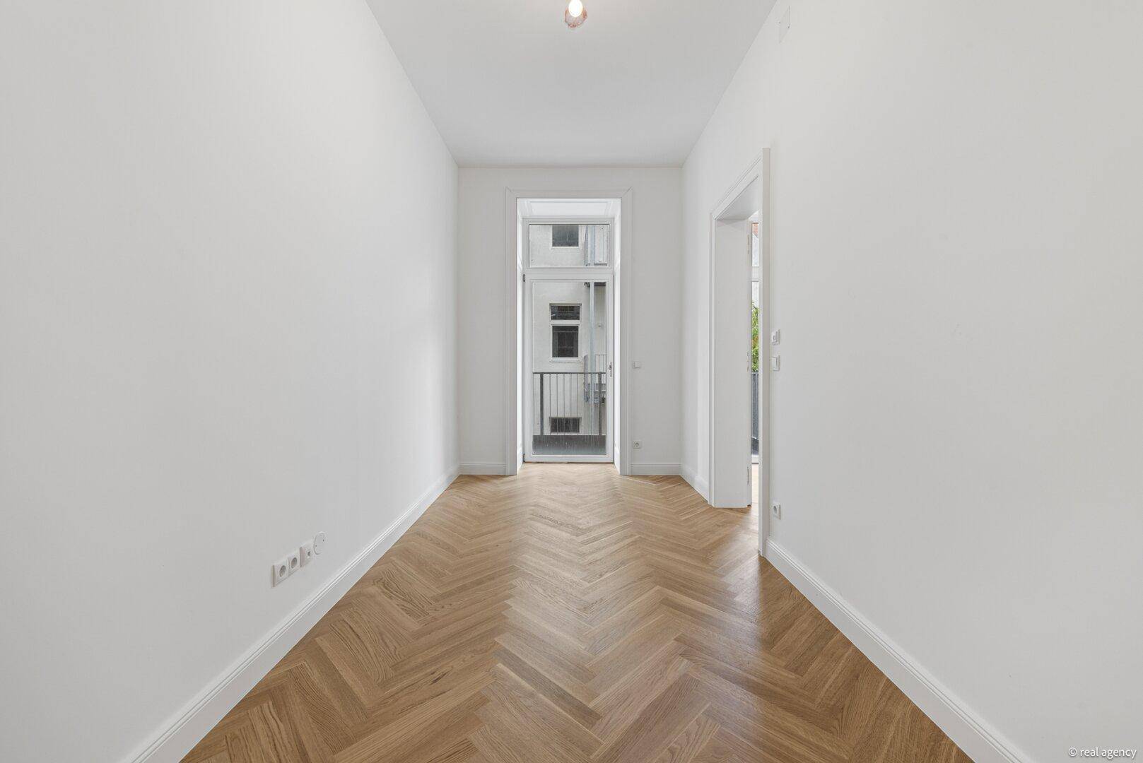 Langer, heller Raum mit Fischgrätparkett und Zugang zu einem französischen Balkon.