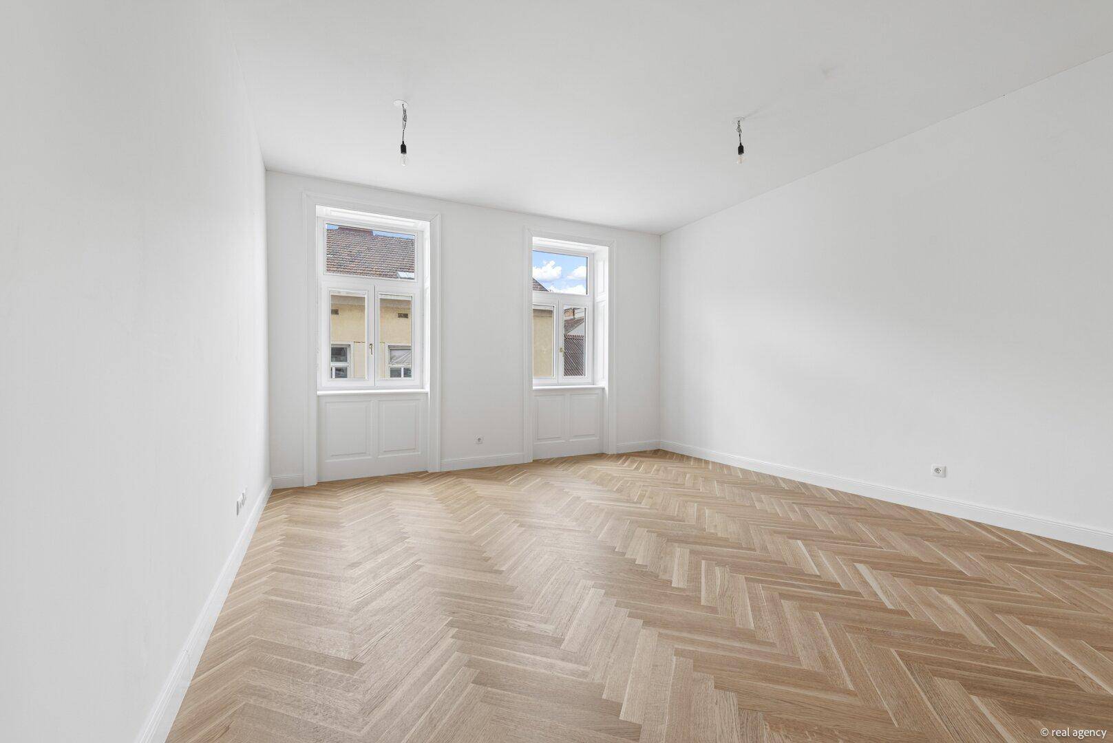 Geräumiger, heller Raum mit Fischgrätparkett und zwei großen Fenstern, bereit zum Einrichten.
