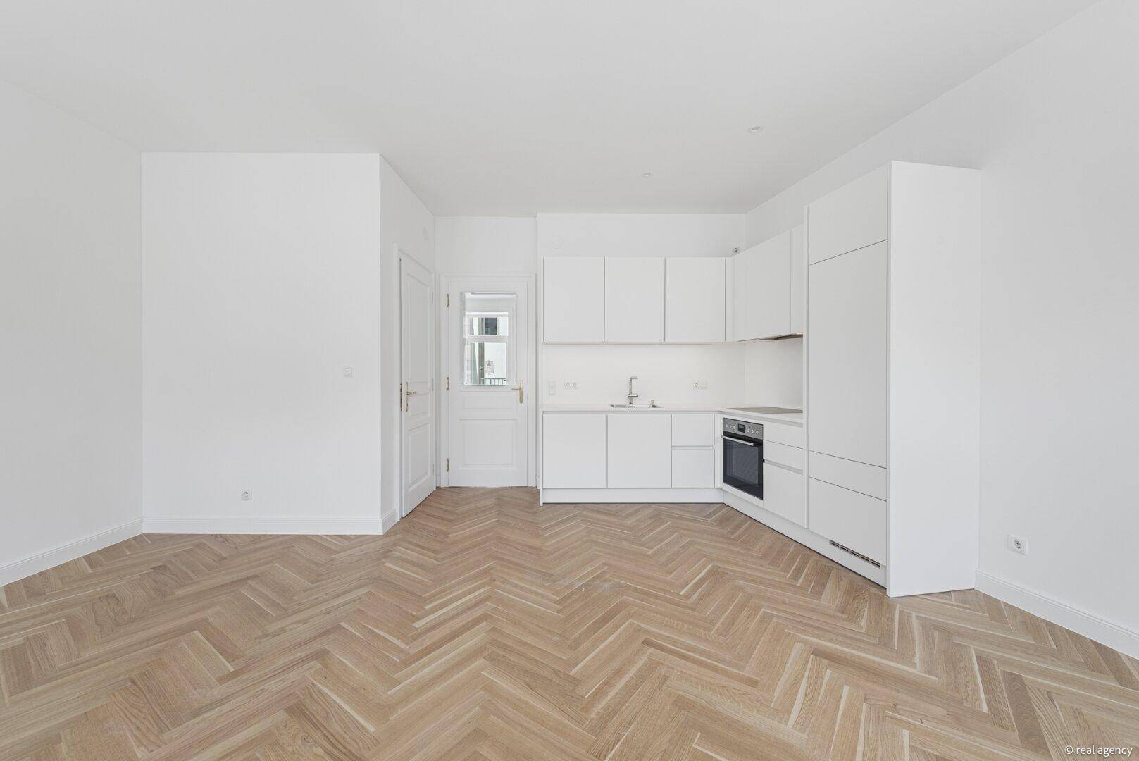 Neue, weiße Einbauküche mit modernen Geräten und Fischgrätparkett in einem hellen Raum.
