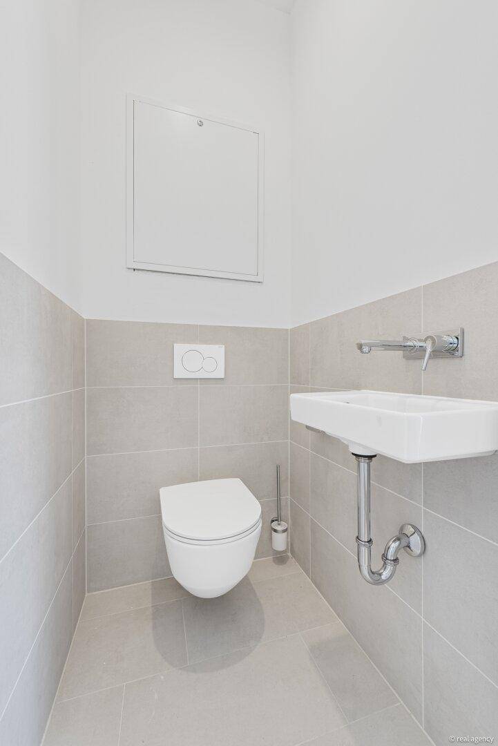 Helles Gäste-WC mit wandhängender Toilette und kleinem Waschbecken, modern gefliest in Grautönen.