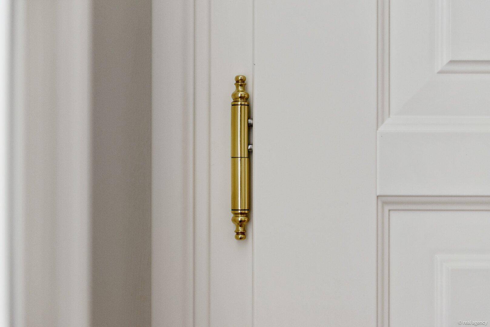 Detailaufnahme eines verzierten goldenen Türscharniers an einer weißen Tür.