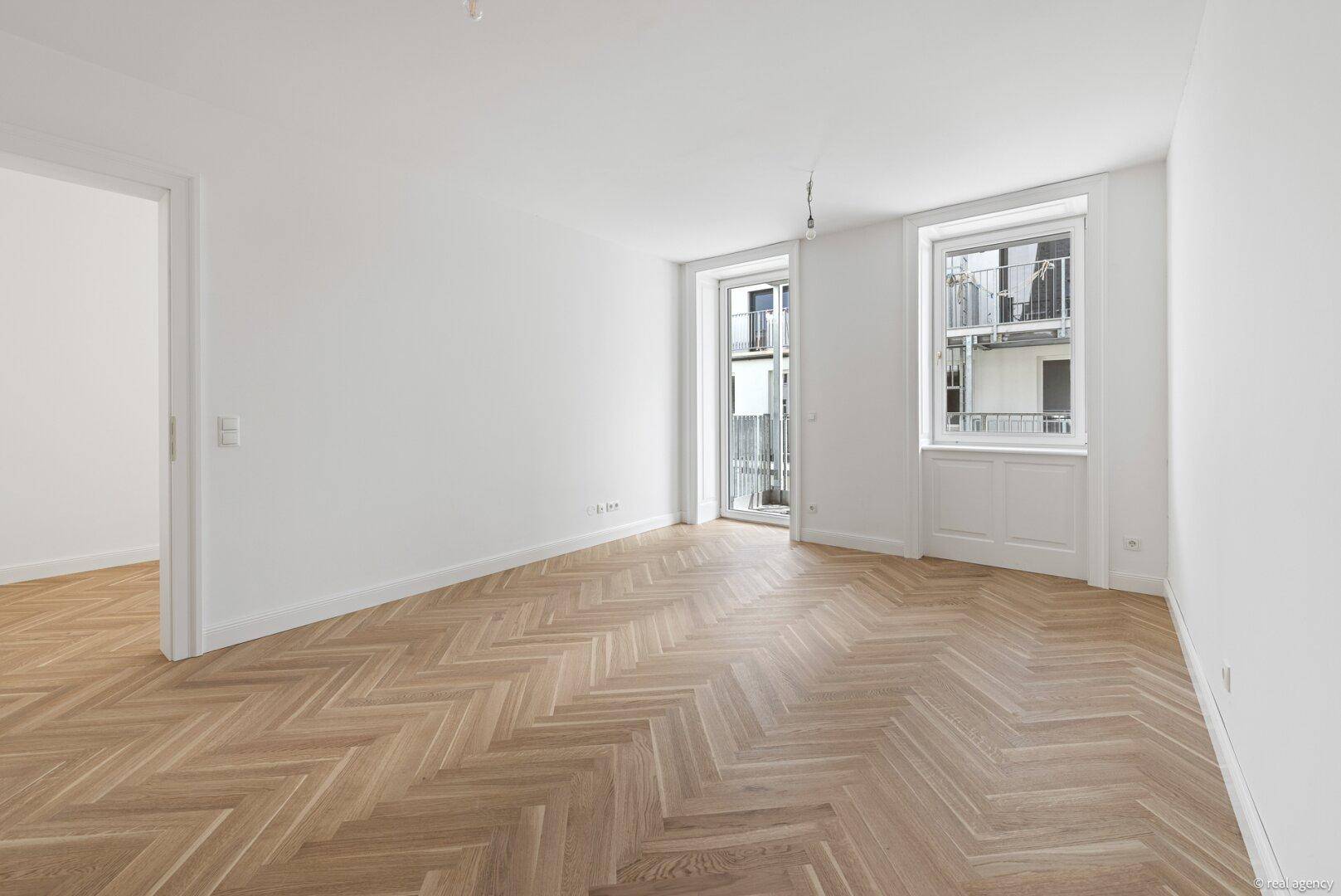 Lichtdurchfluteter Raum mit Fischgrätparkett und zwei Fenstern, die auf einen Balkon führen.